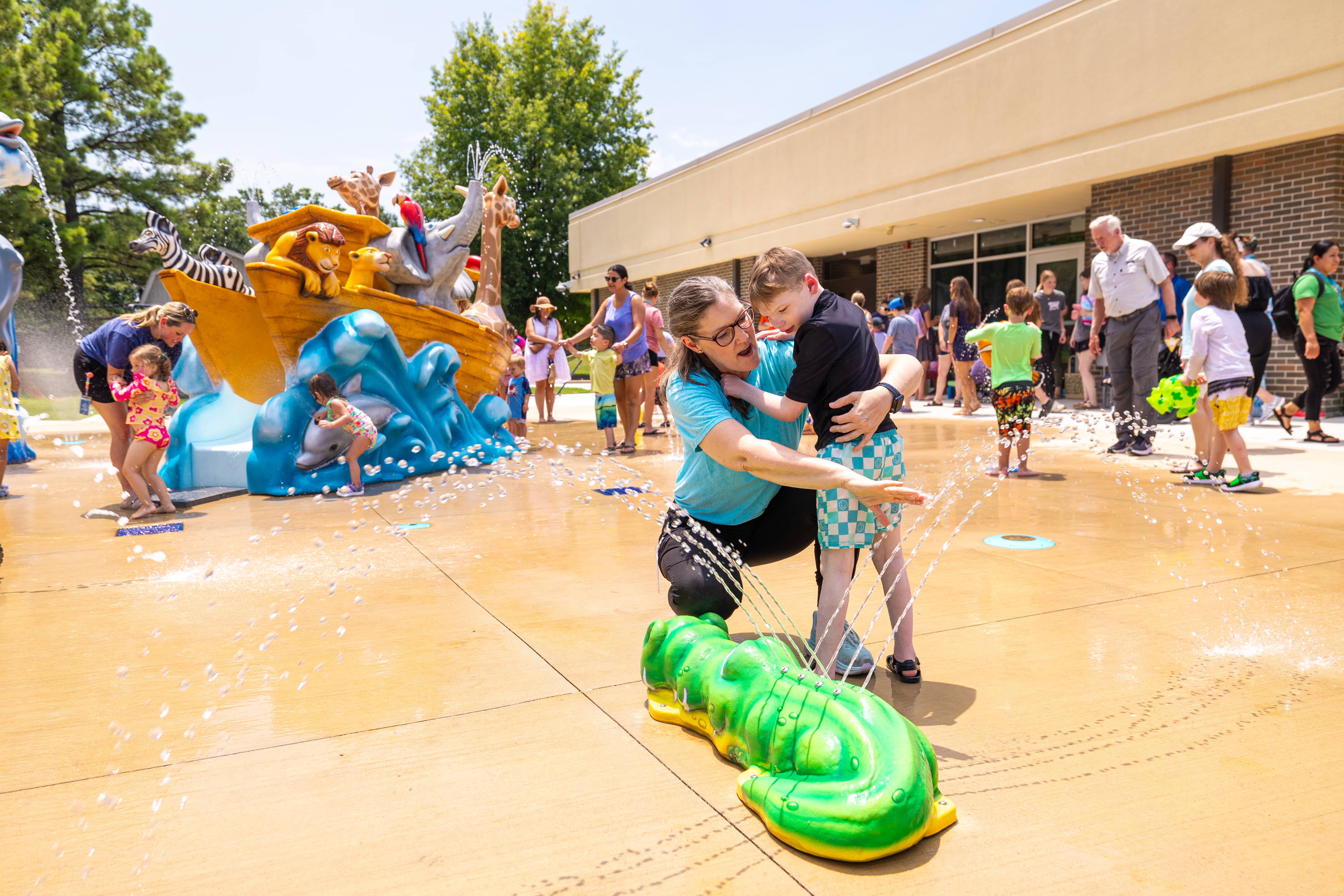 a child and caregiver interacting with an alligator-themed spray feature on a splash pad